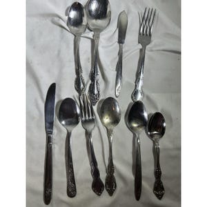 May include: An assortment of vintage silverware, including spoons, forks, and a knife, arranged on a white surface. The silverware has ornate handle designs and a shiny, reflective surface, suggesting a classic or antique style.