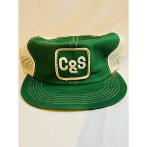 May include: Green and white trucker hat with the letters "C&S" in a white square patch. The hat has a green front panel and brim, with a white mesh back. The brim has white stitching. The hat is a classic style.