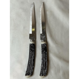 May include: Two vintage steak knives with stainless steel blades and dark brown, textured handles. The handles have a cream-colored base and are curved for a comfortable grip. The knives are likely from the mid-20th century.