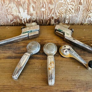 May include: An assortment of vintage car door handles and window cranks. The handles are chrome with some green accents, and the window cranks are silver with black knobs. The items are arranged on a wooden surface.