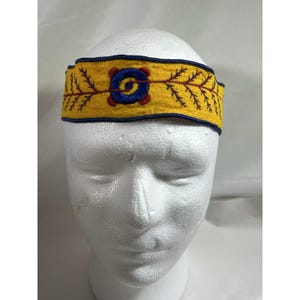 May include: A yellow fabric headband featuring a central blue circular embroidered motif with red accents, flanked by symmetrical red branch-like embroidery patterns. The headband is finished with a dark blue stitched border along the top and bottom edges.