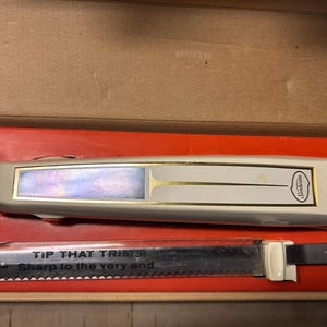 May include: An off-white Sunbeam electric carving knife set in its original box. The knife handle has gold accents and a pearlescent panel. The serrated blade is visible below the handle, with the text "TIP THAT TRIMS Sharp to the very end."