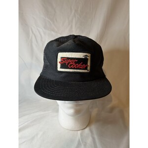 May include: Black trucker hat with a curved brim. The hat features a rectangular patch with the words "Super Cooker" in red script. The patch is outlined in white and has a black background. The hat has a mesh back.
