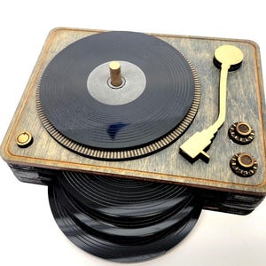 May include: Wooden turntable-shaped coaster set. The coaster set features a gray wooden base with a black record design. The set includes a stack of black record coasters and gold-colored accents. A unique and stylish addition to any home.