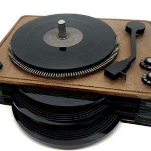May include: A wooden turntable-shaped coaster set. The top features a black record and a tonearm. The base holds several black record-shaped coasters. The wood has a light brown finish.