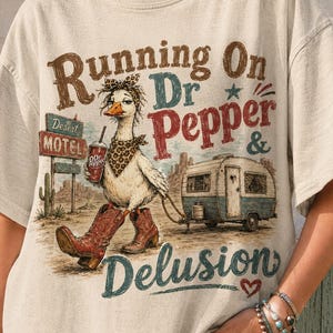 May include: Beige t-shirt featuring a whimsical illustration. A goose wearing a bandana, cowboy boots, and a Dr. Pepper cup is depicted with a trailer. Text reads "Running On Dr Pepper & Delusion" with a desert motel sign.
