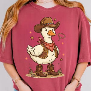 May include: A maroon t-shirt with a cartoon duck in cowboy attire. The duck is depicted wearing a brown cowboy hat, vest, and boots, and a red bandana. The image includes the text "PNG FILE ONLY" at the bottom.