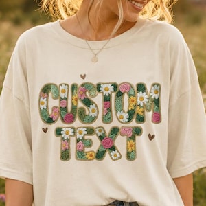 May include: A cream-colored short-sleeve t-shirt featuring the words CUSTOM TEXT in a bold, blocky font. The letters are filled with a colorful floral pattern including daisies, pink roses, and green leaves. The shirt is paired with blue denim shorts.