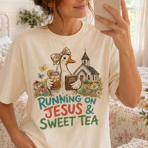 May include: A cream-colored short-sleeve t-shirt featuring a graphic of a duck wearing a floral bow, holding a glass of tea and a book labeled Holy Bible, with a church and flowers. The text reads Running on Jesus & Sweet Tea.