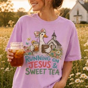 May include: A light purple t-shirt featuring a graphic of a duck wearing a floral bow, holding a mason jar of iced tea and a Holy Bible, with a church and flowers in the background. The text reads, Running on Jesus & Sweet Tea.