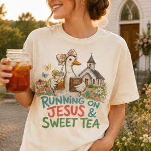 May include: A cream-colored t-shirt featuring a graphic of a duck wearing a floral bow, holding a glass of iced tea and a Holy Bible, standing near a church. The text reads RUNNING ON JESUS & SWEET TEA in green and red lettering.