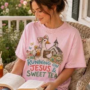 May include: A light pink t-shirt featuring a graphic of a duck wearing a floral bow, holding a glass of iced tea and a book labeled Holy Bible, next to a church. The text reads Running on Jesus & Sweet Tea in teal and red lettering.