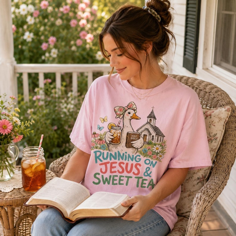 May include: A light pink t-shirt featuring a graphic of a duck wearing a floral bow, holding a glass of iced tea and a book labeled Holy Bible, next to a church. The text reads Running on Jesus & Sweet Tea in teal and red lettering.