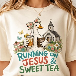May include: A cream-colored t-shirt featuring a graphic of a duck wearing a floral bow, holding a mason jar of tea and a Holy Bible, standing near a church and flowers. The text reads RUNNING ON JESUS & SWEET TEA in teal and red lettering.