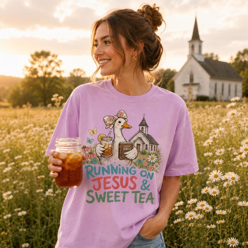 May include: A light purple t-shirt featuring a graphic of a duck wearing a floral bow, holding a mason jar of iced tea and a Holy Bible, with a church and flowers in the background. The text reads, Running on Jesus & Sweet Tea.