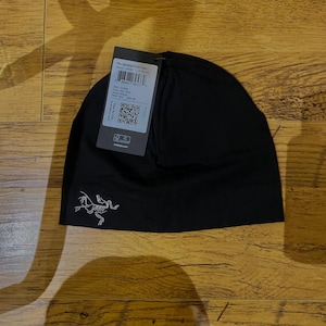 Op de afbeelding: Zwarte beanie met een klein wit logo aan de voorkant. Een productlabel is aan de beanie bevestigd. De beanie is gemaakt van een zacht materiaal en is waarschijnlijk ontworpen voor buitenactiviteiten.