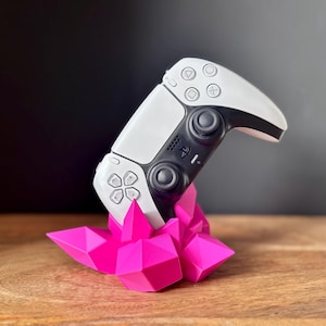 May include: A white and black game controller sits on a bright pink, geometric crystal-shaped stand. The controller features buttons and joysticks. The stand is made of multiple triangular facets.