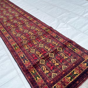 May include: A long, rectangular rug with a detailed geometric pattern in shades of burgundy, red, gold, and navy blue. The rug has a fringed edge and is likely used as a runner. The design features repeating motifs.