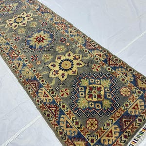 May include: A rectangular area rug featuring a gray background with a repeating pattern of geometric floral medallions in shades of blue, red, yellow, and cream. The rug has a decorative border and short white fringe along the narrow ends.