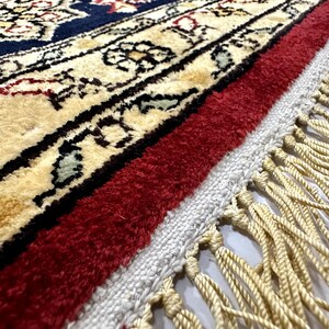 May include: Close-up of a detailed area rug with a cream-colored central design, bordered by a deep navy blue and red. The rug features intricate floral patterns and a fringe of cream-colored tassels.