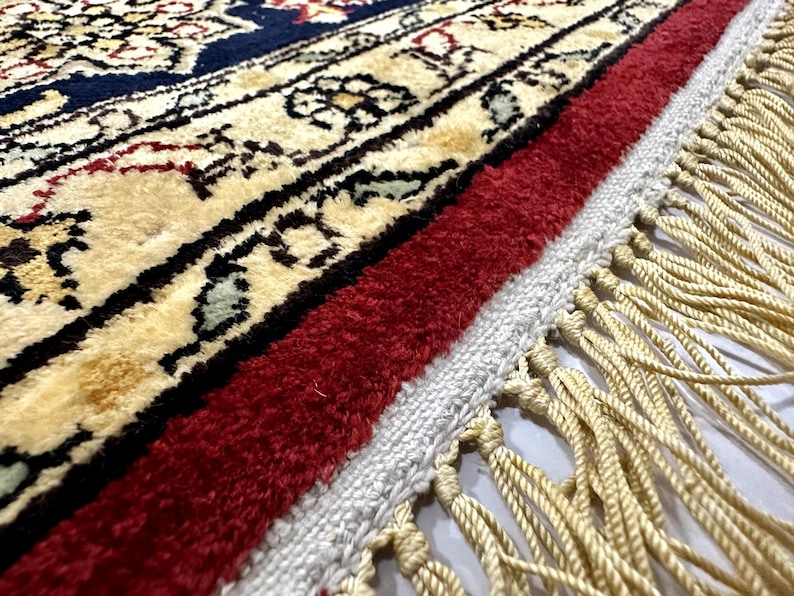 May include: Close-up of a detailed area rug with a cream-colored central design, bordered by a deep navy blue and red. The rug features intricate floral patterns and a fringe of cream-colored tassels.