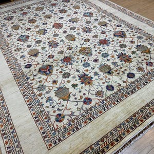 May include: A large, rectangular ivory rug with an elaborate floral design. The rug displays a central field of flowers in blue, red, and gold, framed by a detailed border with geometric and floral patterns. The rug is placed on a wooden floor.