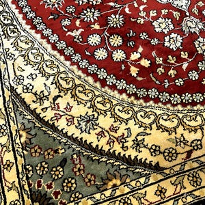 May include: Close-up of a detailed area rug with a rich color palette. The rug features a central red field with white floral patterns, bordered by cream and black sections with intricate designs. The rug's design is ornate and traditional.