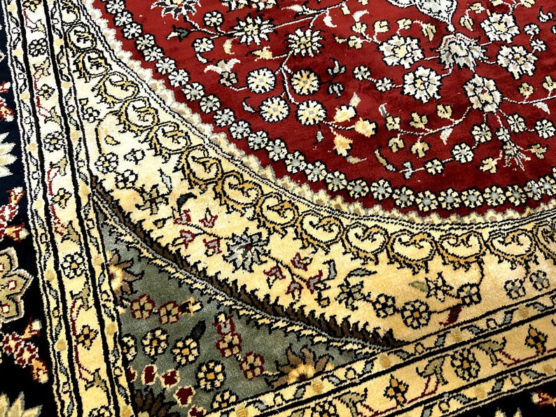 May include: Close-up of a detailed area rug with a rich color palette. The rug features a central red field with white floral patterns, bordered by cream and black sections with intricate designs. The rug's design is ornate and traditional.