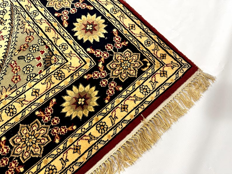 May include: Close-up of a section of an ornate rug with a cream, black, and burgundy color scheme. The rug features floral and geometric patterns, with a fringed edge. The design includes stylized flowers and intricate borders.
