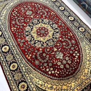 May include: A large, ornate rug with a central oval design in deep red, featuring floral patterns and a cream-colored border. The border includes black and gold floral medallions. Rolled rugs are visible in the background.