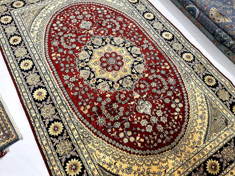 May include: A large, ornate rug with a central oval design in deep red, featuring floral patterns and a cream-colored border. The border includes black and gold floral medallions. Rolled rugs are visible in the background.