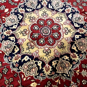 May include: Close-up of a red, beige, and navy blue patterned rug. The rug features a central medallion design with floral and geometric motifs. The border is a deep red with smaller floral patterns.