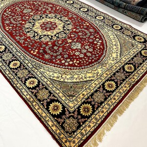 May include: An ornate, rectangular rug with a central oval design. The rug features a deep red field with floral patterns, a cream-colored border, and a black outer border with gold floral accents. The rug has a fringe along the edges.