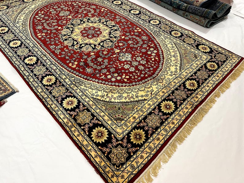 May include: An ornate, rectangular rug with a central oval design. The rug features a deep red field with floral patterns, a cream-colored border, and a black outer border with gold floral accents. The rug has a fringe along the edges.