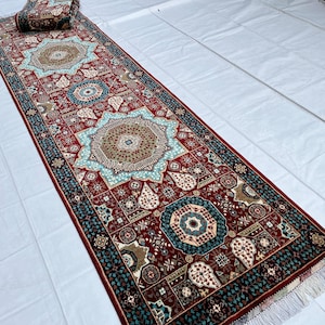 Puede incluir: Una alfombra larga y rectangular con un detallado patrón geométrico en tonos rojos, turquesa y marrón. La alfombra presenta un diseño de medallón central y un borde con flecos. Una sección enrollada de la alfombra es visible en la parte superior.