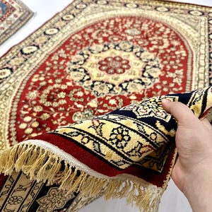 May include: A detailed, ornate rug with a central red field and cream-colored border. The rug features a floral pattern with navy blue and gold accents. The fringe is a light tan color. The rug is partially rolled up, revealing the underside.