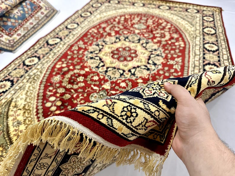 May include: A detailed, ornate rug with a central red field and cream-colored border. The rug features a floral pattern with navy blue and gold accents. The fringe is a light tan color. The rug is partially rolled up, revealing the underside.