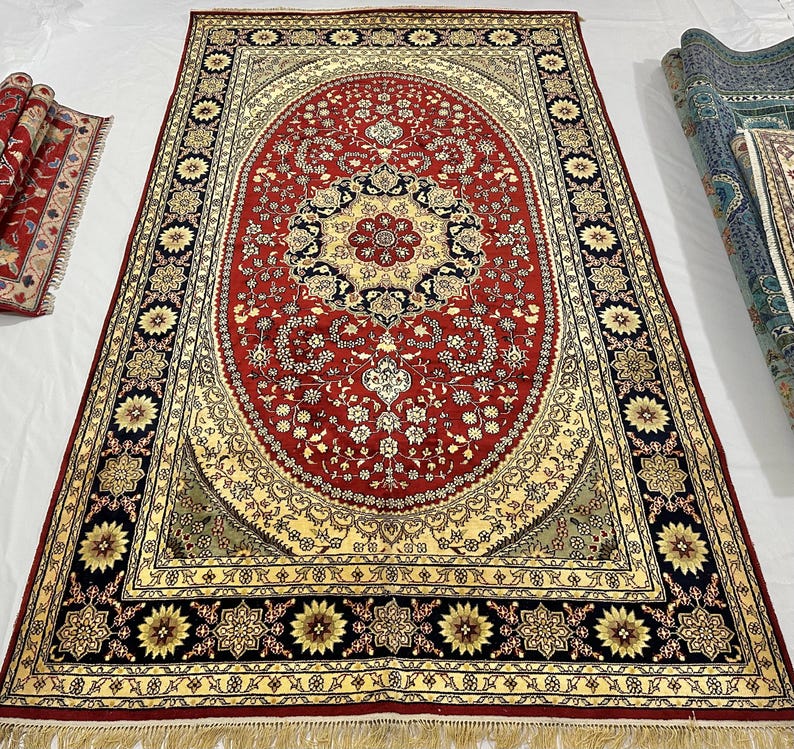 May include: A large, rectangular red rug with an oval central design. The rug features intricate floral patterns in cream, black, and gold. The border has a repeating floral motif with a gold fringe along the bottom edge. A rolled-up rug is visible in the background.