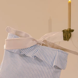 May include: A blue and white striped fabric basket with a white bow. The basket has a ruffled edge and is tied with a large bow. The background includes a candle holder with a lit candle.