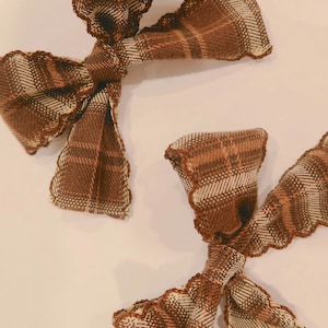 May include: Two hair bows made from a brown, tan, and cream plaid fabric. Each bow features a knotted center and edges finished with a decorative scalloped stitch in a dark brown thread, creating a textured appearance on the fabric surface.
