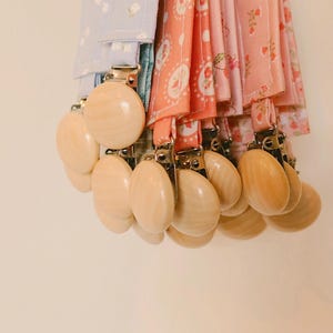 May include: A collection of baby pacifier clips with wooden beads and fabric straps. The fabric patterns include floral and polka dot designs in shades of pink, peach, and blue. The clips are attached to a metal clasp.