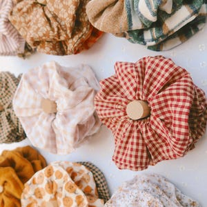 May include: A collection of fabric hair scrunchies in various patterns and colors, including plaid, gingham, floral, and pumpkin prints, each centered on a small, light-colored wooden dowel arranged on a flat surface.