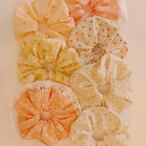 May include: A collection of fabric scrunchies in various floral patterns and colors, including yellow, peach, and cream. Some scrunchies feature delicate lace trim. These hair accessories are perfect for adding a touch of vintage charm to any hairstyle.