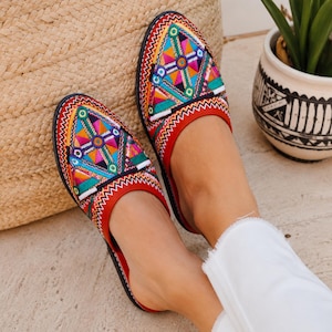 May include: A pair of red slip-on shoes featuring intricate geometric embroidery in vibrant shades of pink, orange, yellow, green, and blue. The pointed-toe footwear has a solid red trim and a black sole, showcasing a detailed triangular pattern.