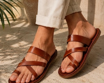 Può includere: Un paio di sandali in pelle marrone con design a infradito, un'ampia fascia orizzontale sul mesopiede e un dettaglio a listini incrociati sul dorso del piede, su una suola piatta con cuciture visibili lungo i bordi.