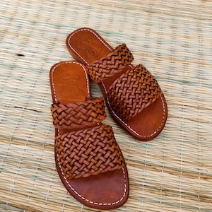 May include: A pair of brown leather sandals with woven straps. The sandals have a flat sole and are stitched with white thread. The sandals are displayed on a woven mat.