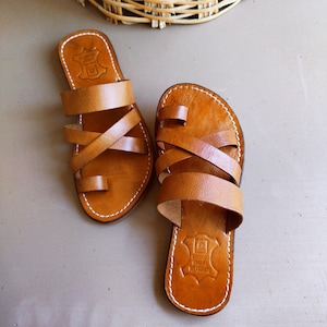 Peut inclure: Paire de sandales en cuir marron clair avec plusieurs lanières et une boucle d'orteil. Les sandales présentent des coutures blanches et un emblème estampé sur la semelle intérieure. Elles sont posées sur une surface neutre.