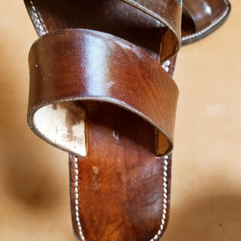Pu&ograve; includere: Primo piano di sandali in pelle marrone con due ampi cinturini. I sandali hanno cuciture bianche sui bordi e una finitura lucida. La pelle sembra usurata, con alcune macchie chiare.