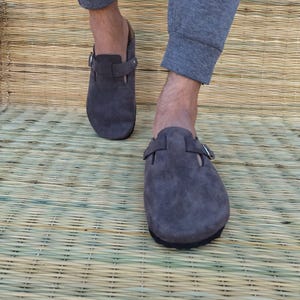 May include: A pair of gray suede clogs with adjustable buckle straps. The clogs have a rounded toe and a black sole. The shoes are shown on a woven mat.