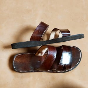 Pu&ograve; includere: Due sandali in pelle marrone con suola scura. I sandali hanno un design semplice con cinturini sulla parte superiore e sono cuciti con filo bianco sui bordi. La pelle ha una finitura lucida.